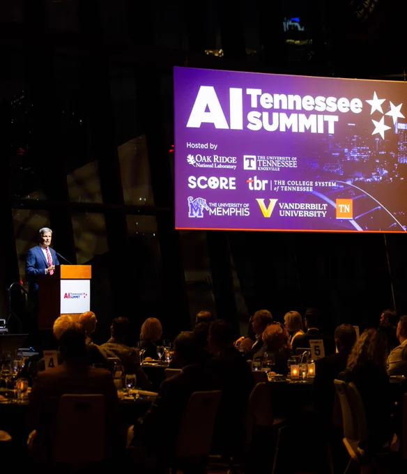 A person, Gov. Biill Lee, speaks at a podium during the AI Tennessee Summit. A large screen displays the event name and logos of sponsoring organizations, including Oak Ridge National Laboratory, The University of Tennessee, SCORE, Tennessee Board of Regents system, The University of Memphis, Vanderbilt University, and the State of Tennessee.  Attendees are seated at tables.