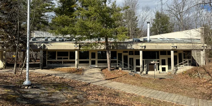 The view of the front of the park office at Frontenac Park after the snow melts. Around the office you can see green pine trees, a leaf covered ground, and a brick-lain walkway that leads down to a lake.