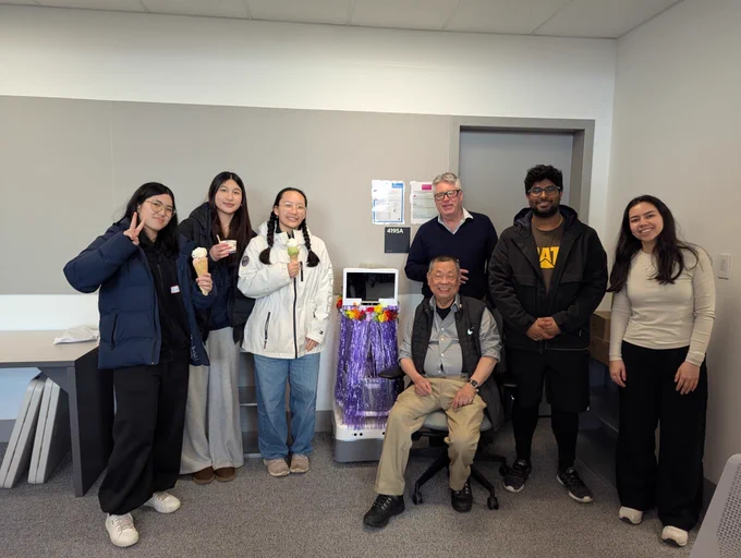 Visiting students, researchers, and partners pose together in the UBC IDEA Lab around the Aether robot, which is decorated with a purple skirt and colourful flowers. The visiting students on the left are holding ice cream.
