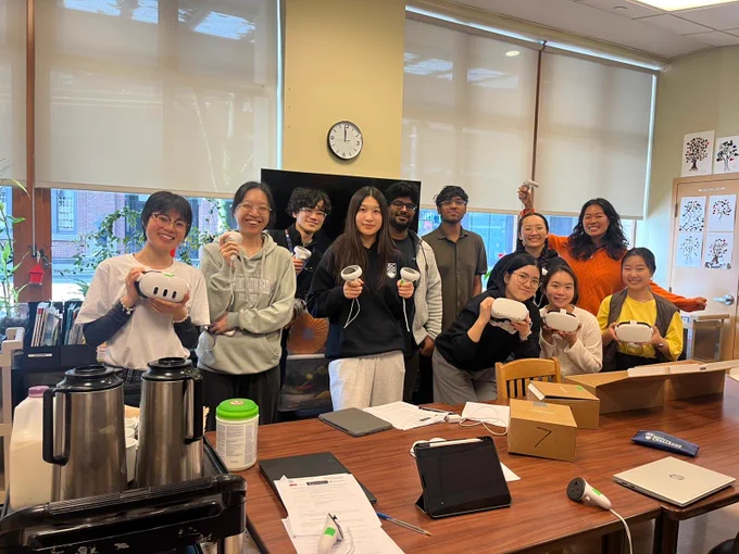 UBC IDEA Lab team posing for a picture at a long-term care site visit, holding VR headsets and controllers, with tables, devices, and project materials visible in the foreground.