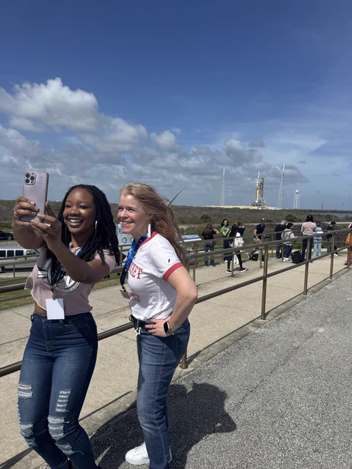 Two people take a selfie with the Artemis II rocket in the background.