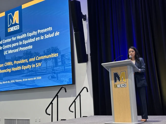 A speaker presents at a UC Merced podium during a Center for Health Equity event, with a screen displaying a presentation on community health and equity.