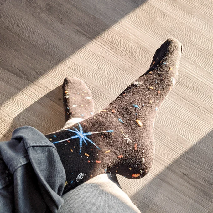Feet crossed on a wooden floor in morning sunlight wearing JWST Deep Field socks, black with pixelated galaxies and a bright blue diffraction spike star