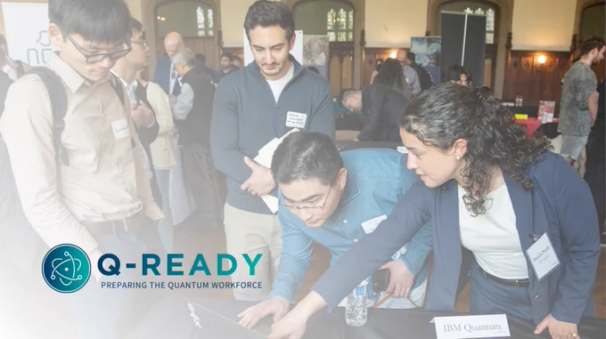 People at an event discussing around a laptop, with a "Q-READY" logo visible in the bottom-left corner.