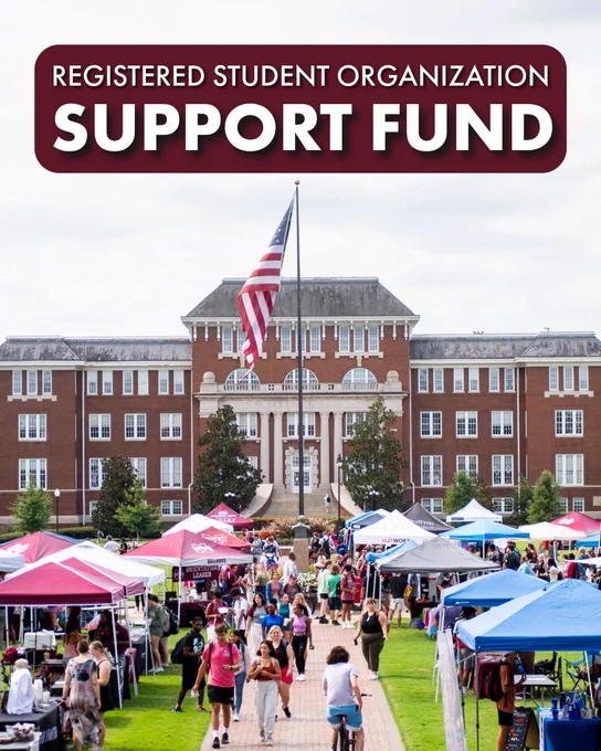 Photo of people walking on the Drill Field sidewalk at Mississippi State with tents set up on each side of the sidewalk. A building and flag pole stand in the background.