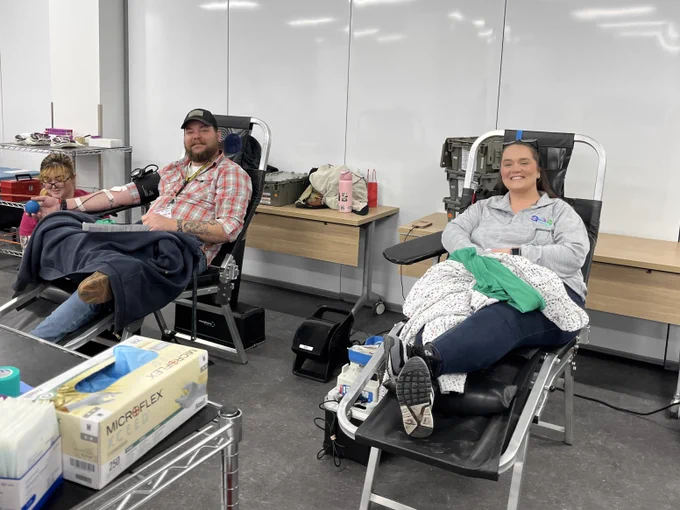Two AES Ohio employees sit in donation chairs during a blood drive, surrounded by supplies and equipment in a large room.
