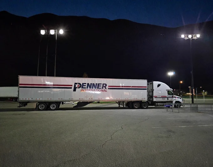 A white Penner International semi truck and trailer parked in a well lit lot at night. The Penner International logo and red and blue stripes are visible on the trailer. Bright overhead lights illuminate the truck while the background remains dark.