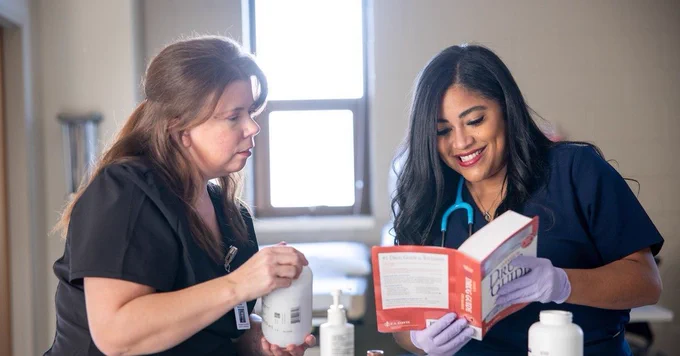 Two healthcare professionals are engaged in a discussion over a book.