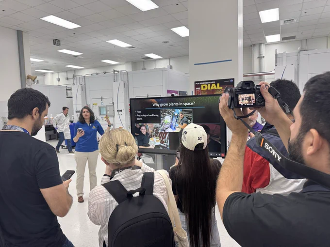 A group of digital creators gathers around a researcher to listen and film as she explains work conducted inside NASA’s plant lab at Kennedy Space Center. They stand in a lab with white walls. A large digital monitor displays photos and information about plant growth in space.