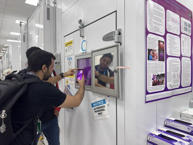 Two people peer into a small square window providing them a view of what’s behind a large closed door. They capture their view on a mobile phone camera. Plant lights emit a purple glow seen through the window.