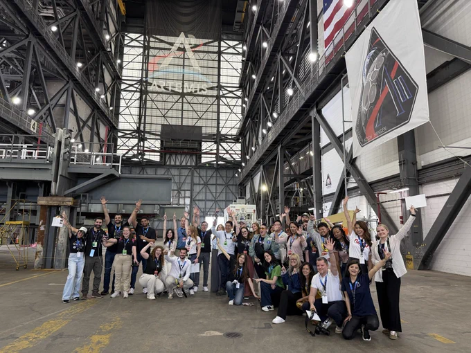 A group poses for a photo inside NASA’s Vehicle Assembly Building at Kennedy Space Center. They are smiling with hands raised in the air.