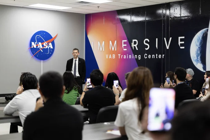 A group of people seated in a classroom-style setting listen to NASA Administrator Jared Isaacman speak. Isaacman is standing at the front of the room wearing a black suit.  Some attendees hold up phones to take photos. NASA and immersive training center graphics cover the back wall.