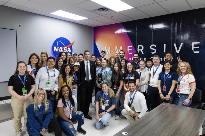 A group of about three dozen people pose together for a group photo. The background features NASA and Immersive Training Center graphics  on the wall.