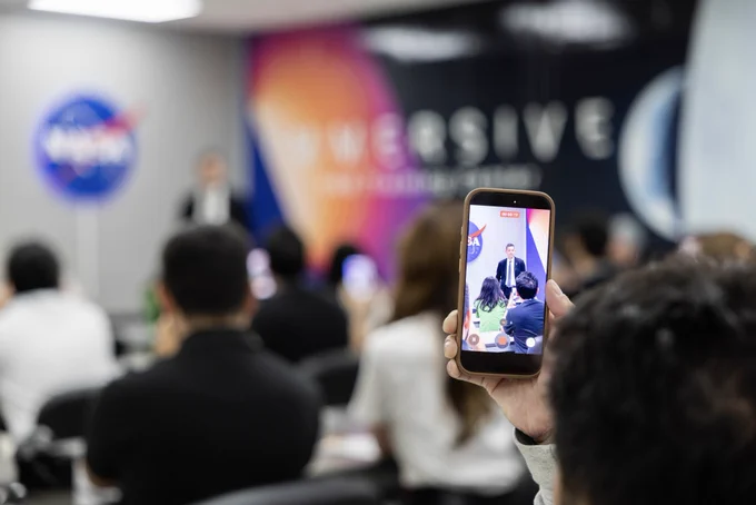 A digital creator seated in an audience at NASA's Kennedy Space Center holds up a smartphone to record NASA Administrator Jared Isaacman as he stands at the front of a room to speak to a group of people.