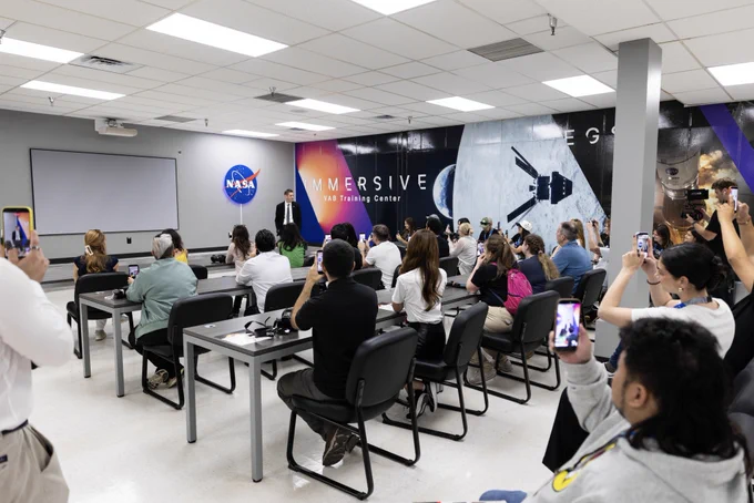 A group of digital creators listen to NASA Administrator Jared Isaacman as he speaks to the group at NASA's Kennedy Space Center. They are seated in a room with rows of tables and black chairs. In the background, there are NASA and Immersive Training Center graphics on the wall.