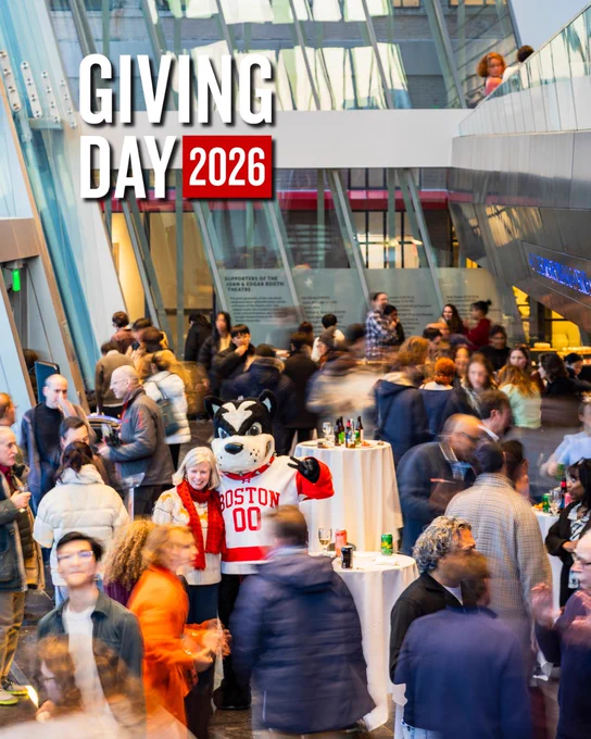 Busy indoor reception in a modern glass-and-steel atrium. A costumed Boston University mascot wearing a red-and-white \"BOSTON 00\" hockey jersey stands near cocktail tables with drinks, smiling and posing for photos. Dozens of blurred guests mingle around the mascot, creating motion blur; some hold plates or glasses. Large white text reading \"GIVING DAY\" with a red \"2026\" box is superimposed across the upper left of the image.