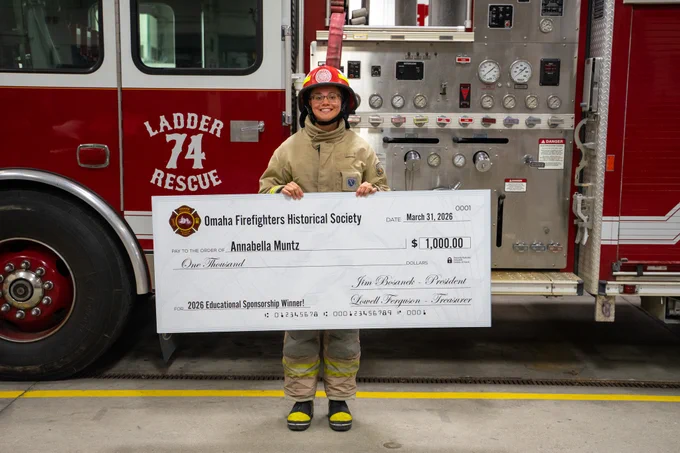 Annabella Muntz posing with an oversized $1,000 scholarship check in front of a firetruck while wearing her firefighting gear.
