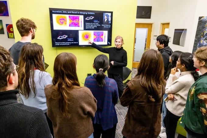 A professor is talking to a group of students while pointing at a television screen in a hallway.