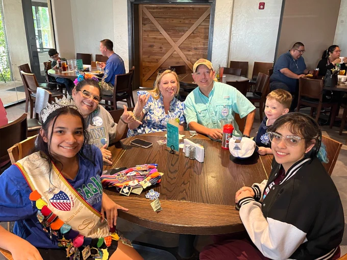 A group of Texas A&M University-Corpus Christi alumni seated around a wooden table in a casual restaurant setting. One individual is wearing a sash with decorative pins, and the table has drinks and a menu on it. The background shows other patrons and a wooden door.