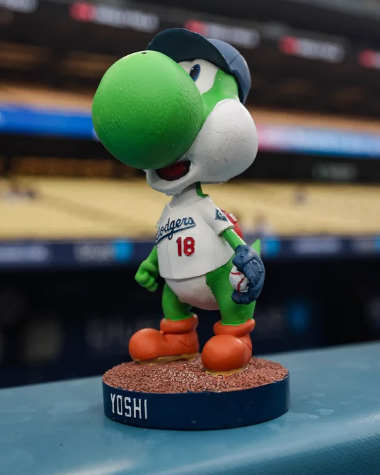 A close-up shot of the Yoshi bobblehead being given away at Dodger Stadium tonight. The bobblehead features Yoshi wearing Yoshinobu Yamamoto's No. 18 Dodgers jersey, a blue Dodgers cap, and a blue glove with a baseball nestled inside the webbing. The placard at the bottom of the bobblehead reads "Yoshi."