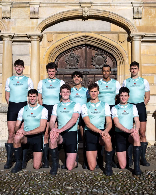 A group of nine individuals wearing matching light blue Cambridge boat team uniforms pose in front of a historic, ornate wooden door set in a stone building. They are positioned in two rows, with some kneeling and others standing. They are all wearing black shorts and boots. The setting appears to be a formal or historic environment.