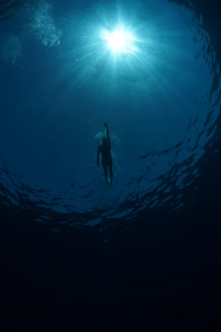 A swimmer (Lewis Pugh) is seen from below in deep blue ocean water, reaching one arm toward bright sunlight at the surface above. The surrounding water appears calm and mostly empty. (Red Sea)