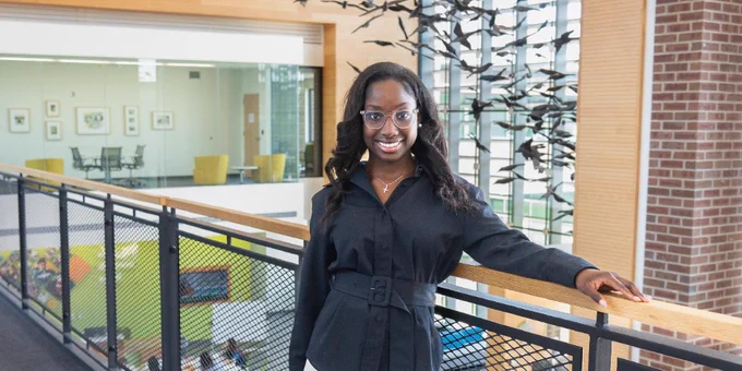 PascalineGrace wearing a black blouse and white trousers on the second floor of the Parkland U-Wing. She leans against a wooden rail with a smile. Black bird sculptures hang behind her in a modern well-lit academic setting. 