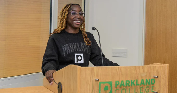 A person with long hair and glasses is speaking at a podium with a microphone. The podium has a "Parkland College" logo. They are wearing a black sweater with "Parkland" printed on it.