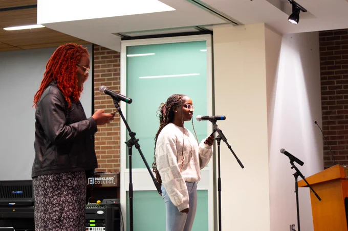 Two people are standing at microphones on a stage in front of a small audience, performing at Parkland. The setting has a modern, well-lit interior with a brick wall and wooden ceiling.