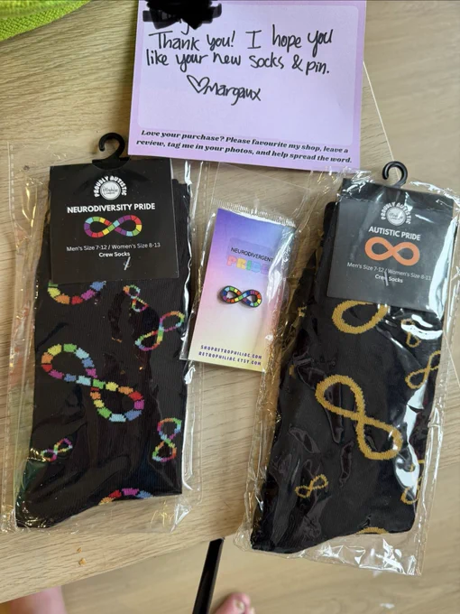 Picture of two pairs of black socks, one with the neurodiversity infinity symbol (rainbow), and the other with the autistic infinity symbol (gold), with a neurodiversity infinity symbol pin between them, and above them a pink card with the name redacted saying "Thank you! I hope you enjoy your socks & pin! Margaux". Yes, @retrophiliac, I *love* them!