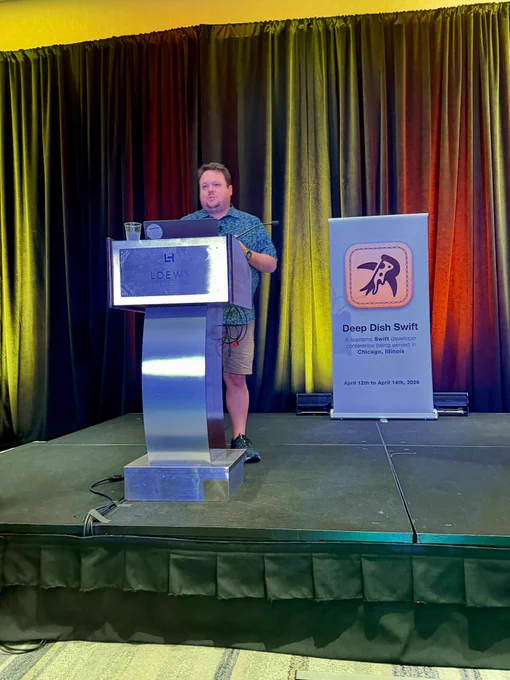 John Marc speaking on a stage, behind a podium and a sign reading "Deep Dish Swift"