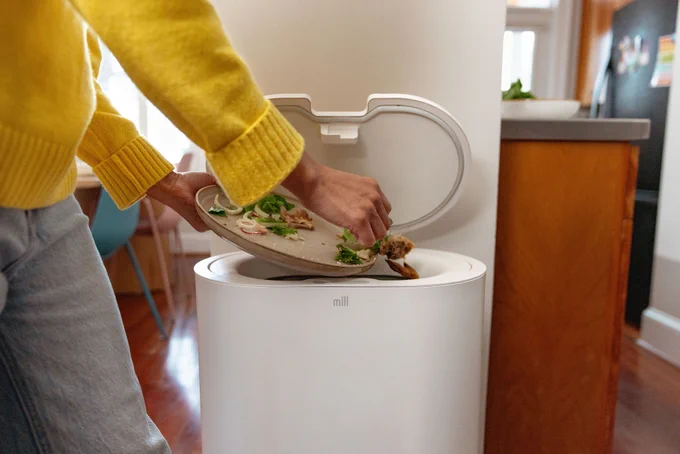A person is scraping food scraps from a plate into a white Mill compost bin in a kitchen setting. 
