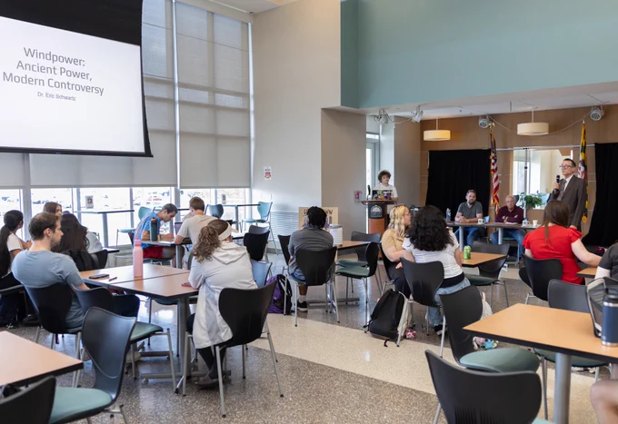 Room full of people seated at tables with projector screen and professor with microphone