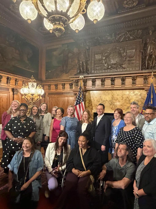 Image of a group of people in an ornately decorated room. The group is visually diverse in race and disability. Advocates, government officials, and DRP CEO Jennifer Garman stand throughout the group with Governor Shapiro at the center. 