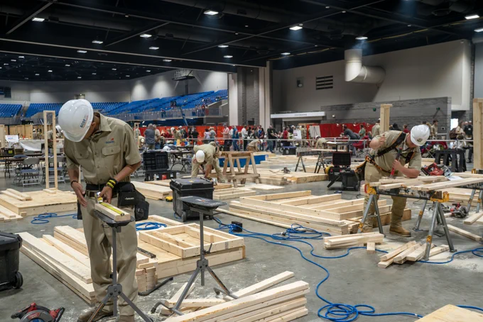 Multiple people working on large wooden structures in a construction site with warehouse-style surroundings.