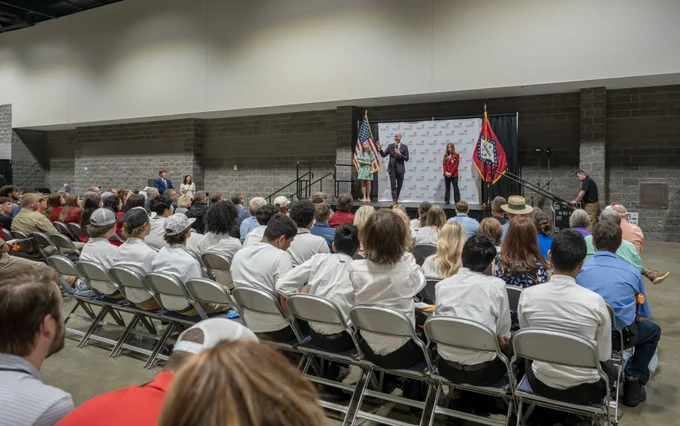 An indoor ceremony with people seated in rows facing a stage, where several people are standing and speaking on stage.