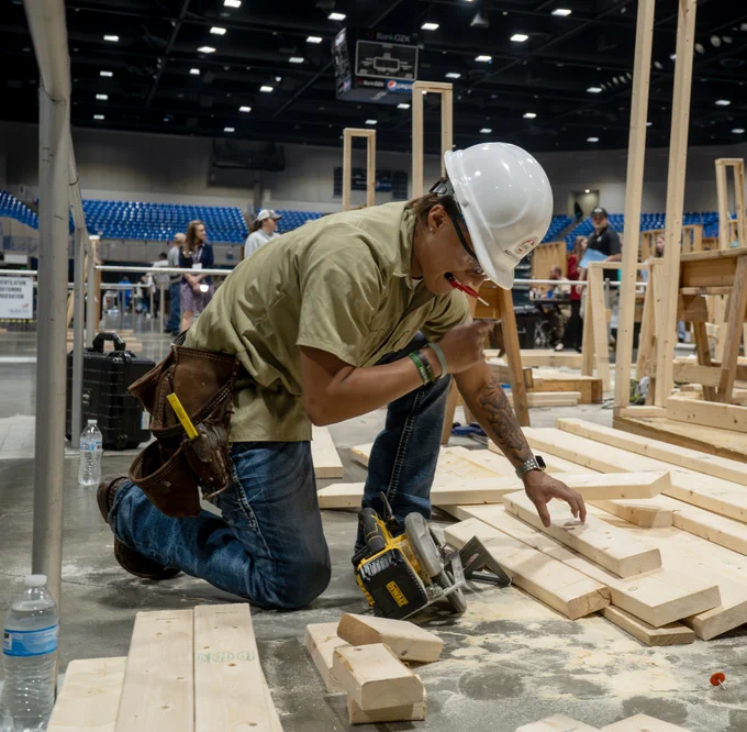 A construction worker wearing a hard hat and jeans, using a power tool to work on wooden planks on the floor of what appears to be a construction site or exhibit hall.