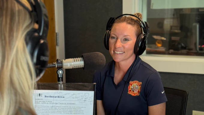 A person wearing a shirt with a fire department logo is sitting in a recording studio, speaking into a microphone. Another individual is seated across, partially visible, also in front of a microphone. Both are wearing headphones.