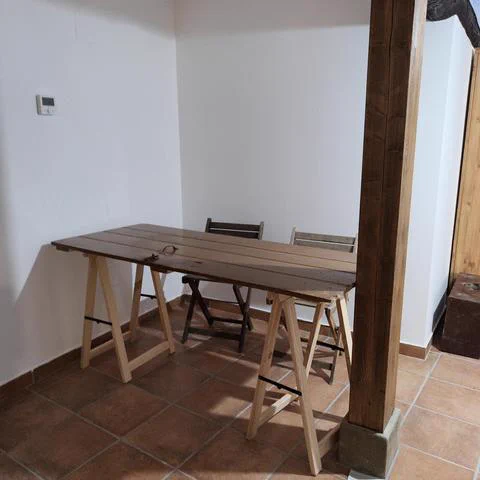 Una puerta de madera puesta sobre dos caballetes a modo de mesa. Encaja justo entre una viga con un pilar de piedra y la pared. Los caballetes son de madera de pino (de color claro), sin pintar  y la puerta está pintada de color marrón oscuro. También hay dos sillas de madera. El suelo es rústico pero el espacio en conjunto se ve arreglado, con las paredes recién pintadas de cal blanca. En una de las paredes se ve un termostato digital.