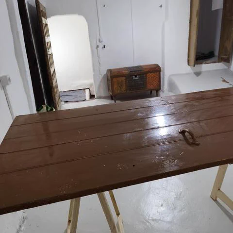 Detalle de la mesa con otra perspectiva. En un primer plano se ve un detalle de la puerta convertida en mesa y al fondo está la puerta de entrada, que es de madera, muy antigua, de tipo castellana. Esta puerta es más alta que el propio hueco de entrada. Junto a ella hay un baúl antiguo chapado en colores rojos y verde oscuro y un poco más allá se observan unas puertas de madera, como si fueran de un armario, pero que en realidad son la entrada a una fresquera. 