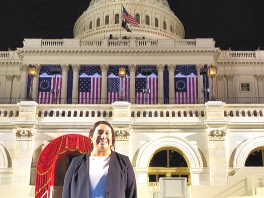 A Texas A&M University-Corpus Christi Islander alumna stands smiling in front of the U.S. Capitol building. The building is adorned with American flags and has a prominent dome. The steps leading up to the building are visible, and the atmosphere suggests a formal event.