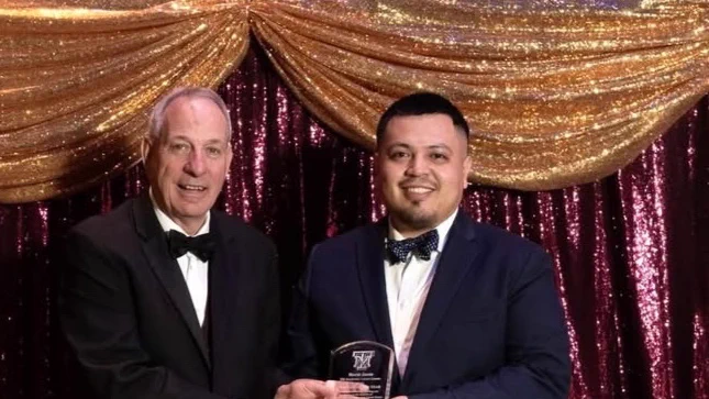 Two individuals in formal attire stand in front of shimmering gold and burgundy drapes. A Texas A&M University-Corpus Christi Islander alumnus is holding an award plaque. Both are smiling at the camera.