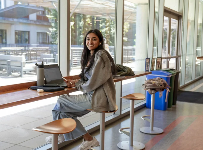 Student smiles while sitting at a long table with a laptop in front of her 
