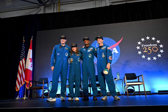 The astronauts of Artemis II (from left: Jeremy Hansen, Christina Koch, Victor Glover, and Reid Wiseman) stand on stage after a crew return event. They are wearing blue space suits and brimmed caps with their names on them. Wiseman is holding Rise, the mascot and zero gravity indicator of the Artemis II mission. Credit: NASA/Helen Arase Vargas