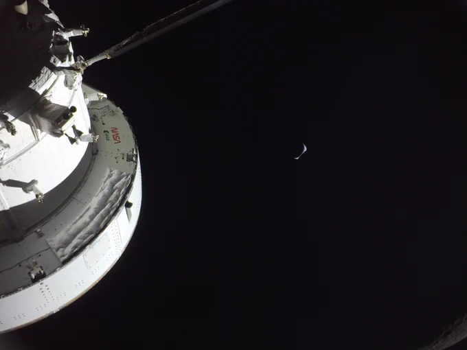 A crescent Earth rises over the horizon of the Moon, which is cutting into Earth's bottom-right edge. The Moon itself is mostly in shadow. The Orion spacecraft is partially visible on the image's far left. Credit: NASA