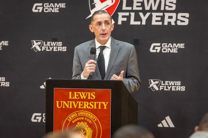 A person stands at a podium with a microphone, speaking at an event. The backdrop features the Lewis University Flyers logo and branding. The podium displays the Lewis University emblem.