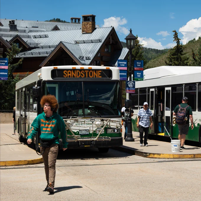 Two Town of Vail buses are parked at a bus station labeled "Sandstone" and "West Vail Green." Several people are walking nearby, with mountains and trees visible in the background. The scene suggests a vibrant, sunny day in a mountainous area.