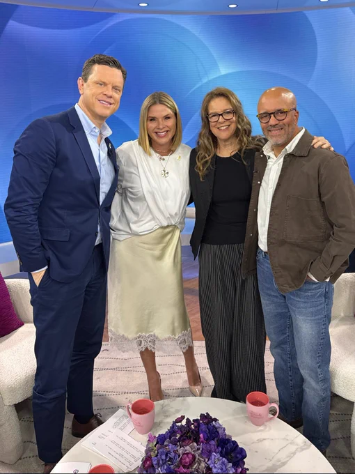 Four people stand together smiling on a TV studio set with bright lighting and a blue backdrop. Carlos Whittaker stands with hosts during a TODAY show segment, posing in a casual group photo on set.