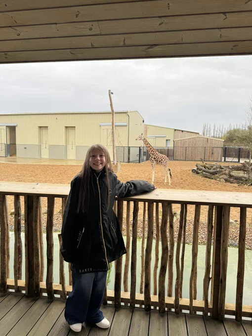 Smiling girl on a wooden viewing platform at a zoo, a giraffe stands in the sandy enclosure behind her.