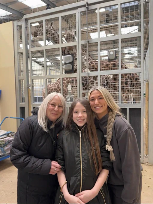 Three smiling people posing in front of a giraffe enclosure inside an indoor animal facility.
